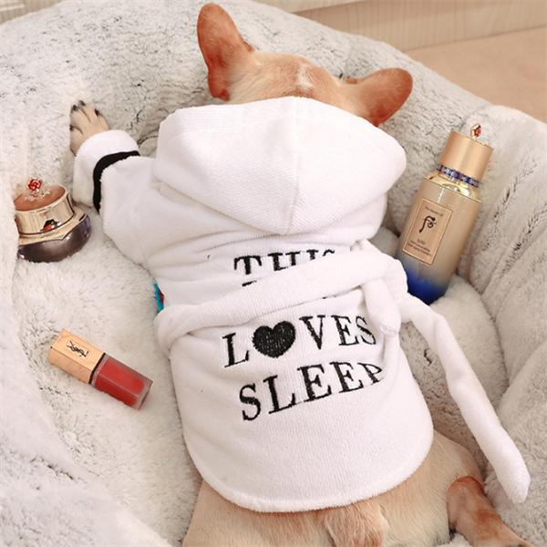 White Hooded Dog Bathrobe – dog wearing a white robe with 'This loves sleep' text, surrounded by beauty products on a fluffy surface